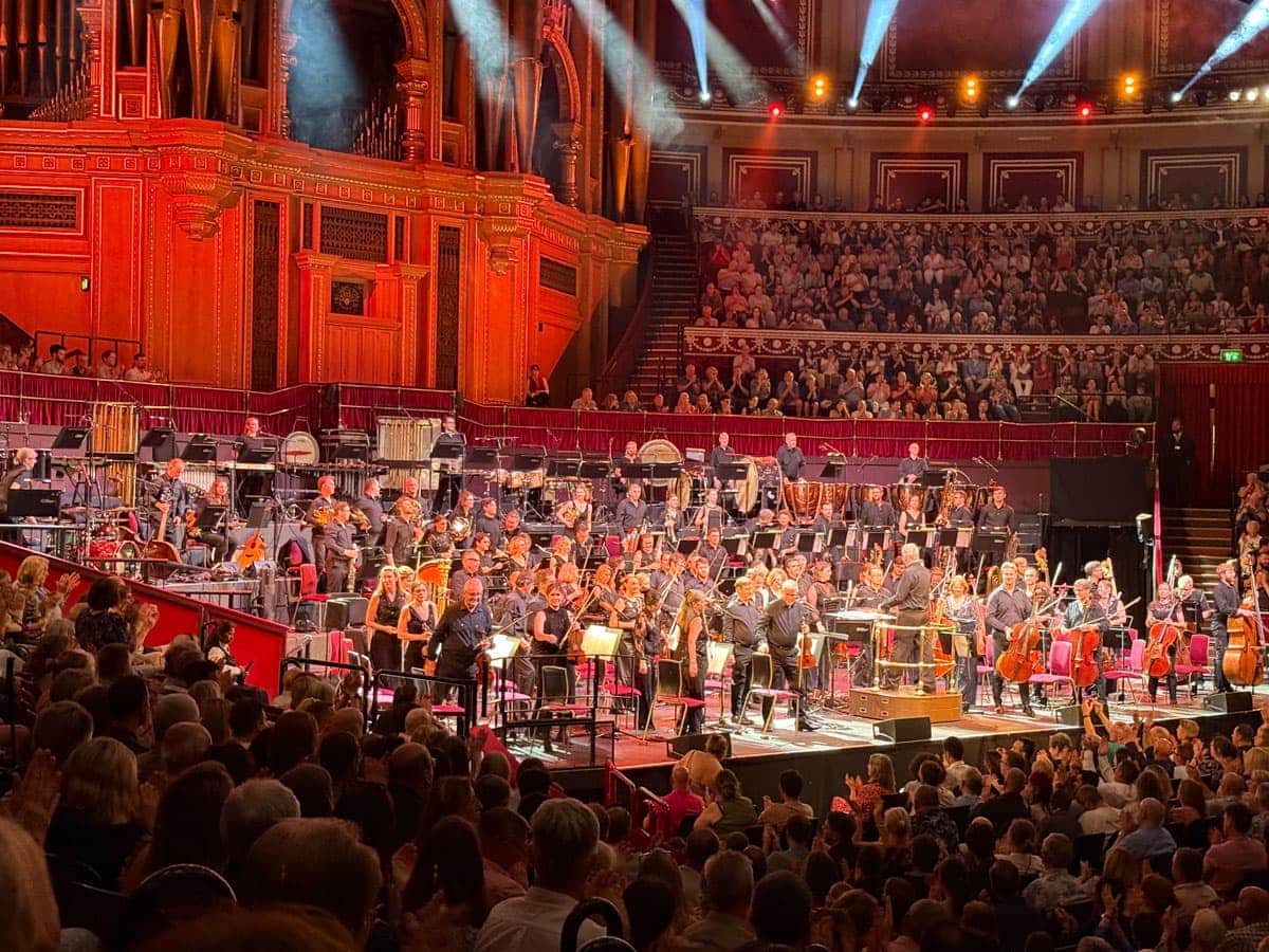 The Royal Philharmonic Orchestra stands for its bow on the Royal Albert Hall stage as the audience applauds, conductor on the podium facing the players. The Royal Philharmonic Orchestra stands for its bow on the Royal Albert Hall stage as the audience applauds, conductor on the podium facing the players.